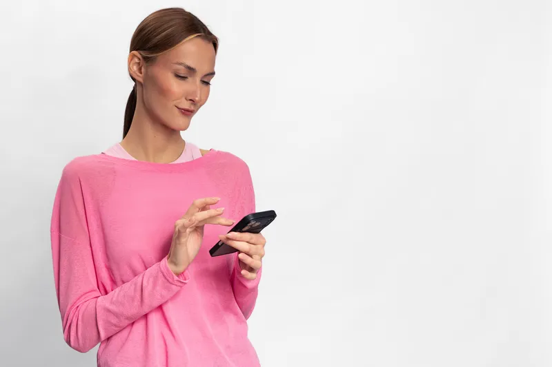 Young woman in pink sweater uses smartphone with focus and slight smile, isolated on white background. Concept of connectivity, technology, communication, and casual lifestyle.