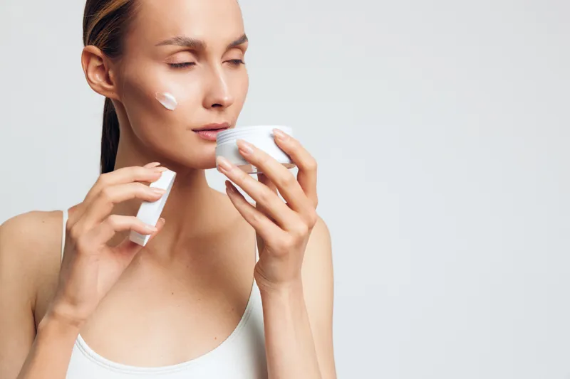Young woman with clear skin applying cream on her face holding a white jar and a small container, eyes closed, soft natural light, minimalistic background, skincare routine concept