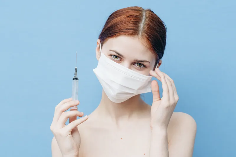 Женщина со шприцем в руке и медицинской маской, делающая инъекцию на синем фоне