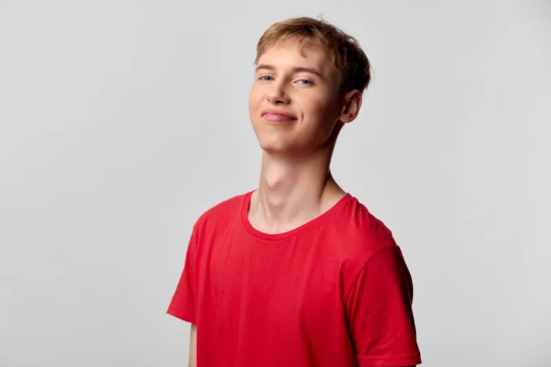 Smiling young man in a red shirt stands against a light gray background, exuding casual confidence, approachable vibe and bright mood ideal for lifestyle and stock photography