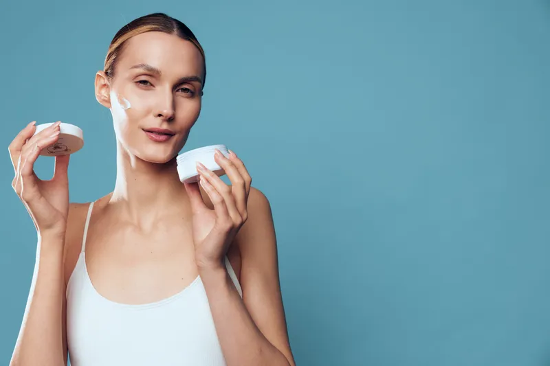 Young woman with blonde hair applies white face cream on her cheeks while holding an open jar against a blue background in a skincare routine.