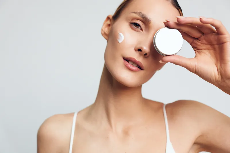 Young woman with clear skin applies moisturizing cream on face holds cosmetic jar near eye looks at camera with soft natural makeup isolated on white background beauty care concept