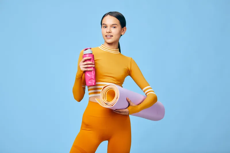 Female fitness enthusiast in bright orange activewear holding a water bottle and a rolled yoga mat against a blue backdrop, ready for a workout, sport training, and healthy lifestyle.