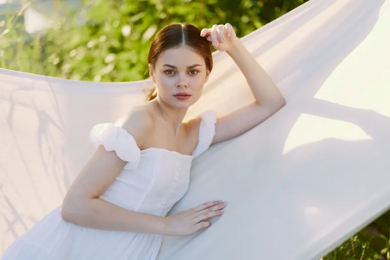 Young woman in a white dress, resting on a hammock in a green garden Fresh atmosphere, bright emotions, and a serene background suggest relaxation and summer vibes