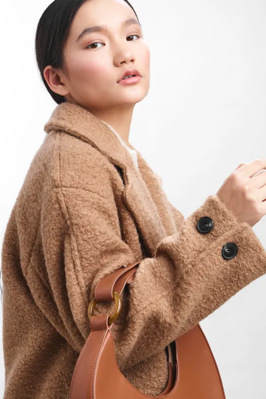 Asian woman wearing a warm coat stands stylishly with a handbag, posing in a bright studio, conveying elegance, confidence, and modern fashion in a composed portrait.