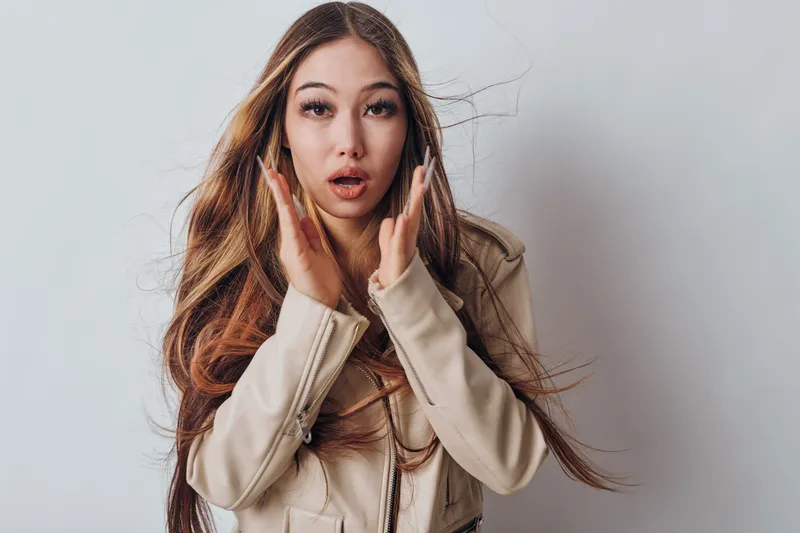 Young woman with long hair and light jacket showing surprise or shock while posing on gray background in studio. Female model and emotion with hands near face and open mouth expression.