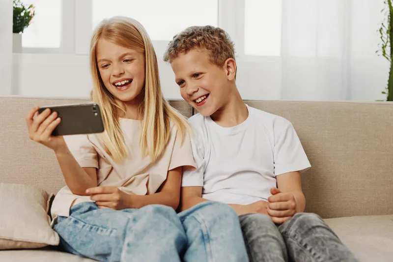 Two happy children sitting on a sofa watching smartphone screen together, smiling and enjoying digital entertainment in bright cozy living room environment.