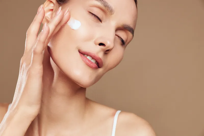 Woman applying skincare cream to face, gentle spa wellness moment, closed eyes and soft smile, hand on cheek, studio beige background, natural light, calm skin glow softly