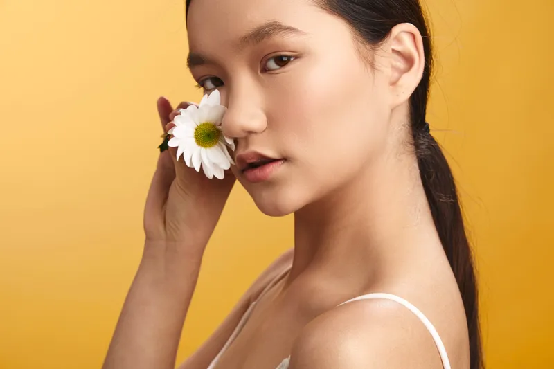 Asian woman holds a white daisy near her face in a bright studio, presenting calm elegance and natural charm with a warm, soft glow and inviting mood.