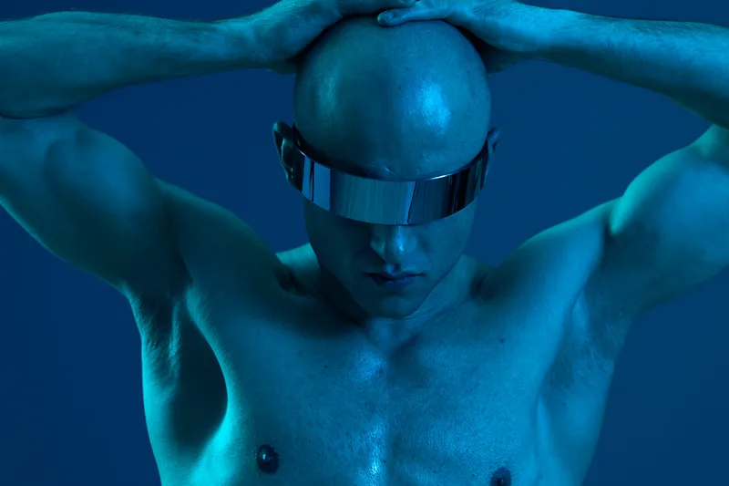 A futuristic man with a bald head, wearing reflective sunglasses, is posing with his hands on his head. The image features a cool color palette emphasizing blue tones.