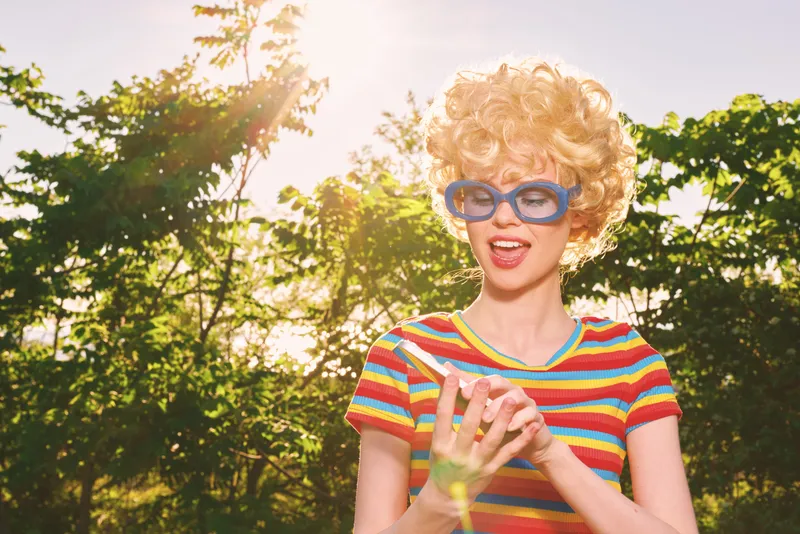 Smiling woman with curly hair wearing a striped shirt and retro sunglasses outdoors under sunlight, casual lifestyle, playful mood, outdoor photography, vibrant summer colors, candid moment.