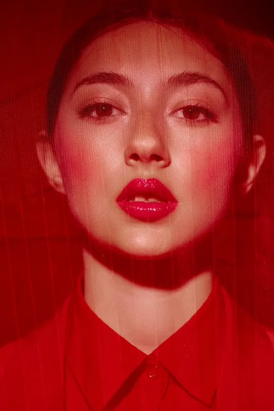 Close-up portrait of a woman wearing glossy red lipstick under a translucent red fabric. Her serious expression and bold makeup create a striking fashion and beauty concept.