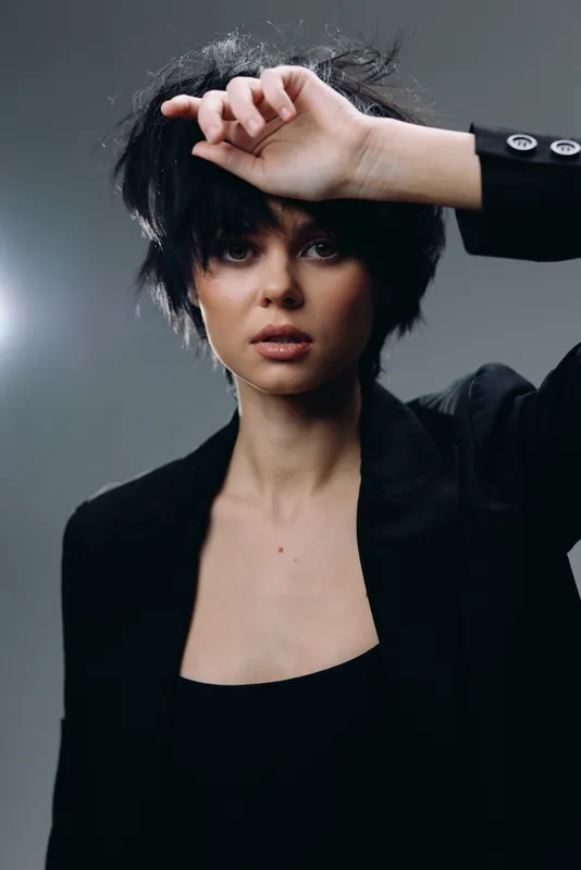 young woman with short black hair wearing black jacket posing confidently with hand near forehead