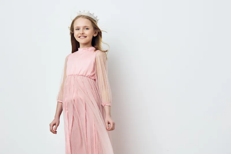 Young girl in a beautiful pink dress with transparent sleeves, wearing a floral crown and smiling joyfully, against a simple white background