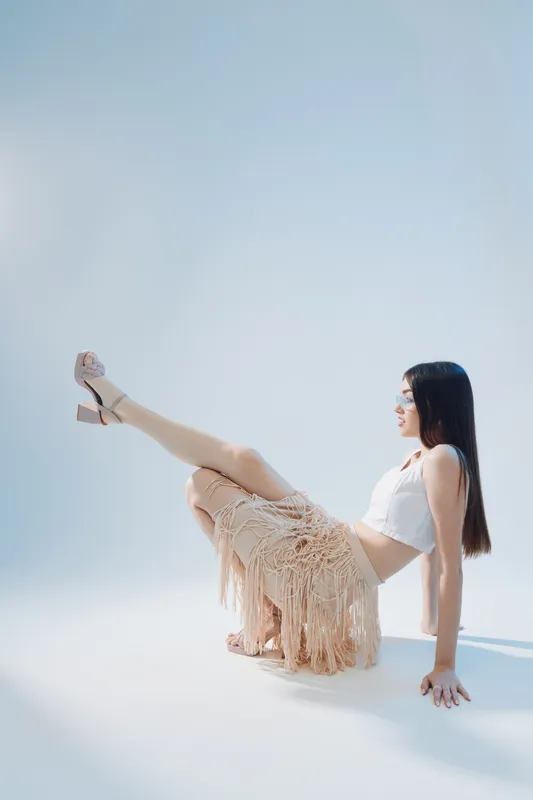 Young woman in a white top and fringed skirt poses on the floor with one leg raised. Studio shot on a light background. Concept of beauty, fashion, and elegance.