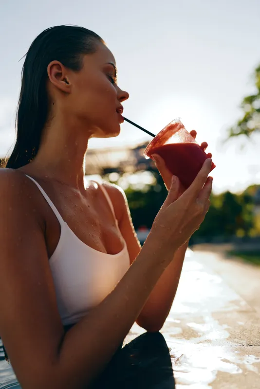 Young woman with wet hair drinks a red smoothie through a straw while relaxing outdoors in a white tank top during sunny weather in a casual lifestyle setting.