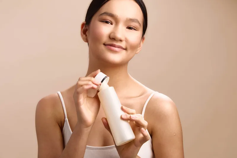 asian woman smiles softly while applying skincare product in a bright minimal space, presenting a calm personal routine that promotes wellness, selfcare, and healthy glow in a modern environment.