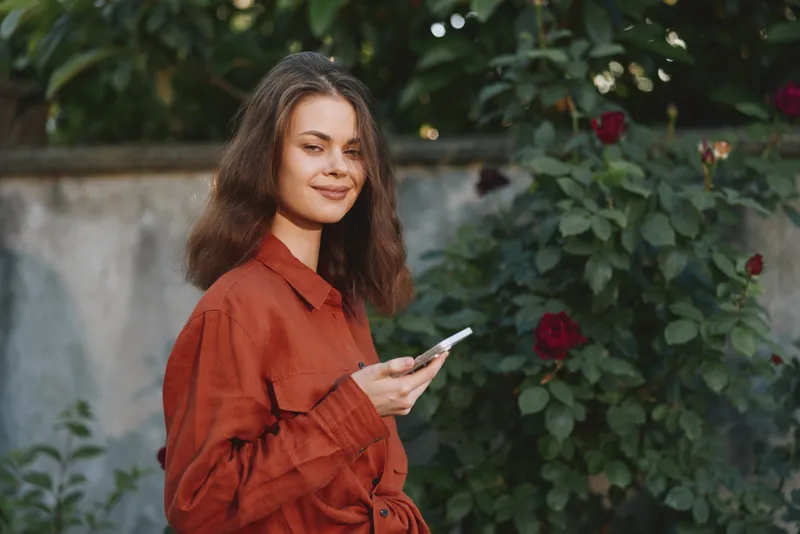 Young woman enjoys a casual outdoor moment, using a smartphone among blooming roses, relaxed mood, warm tones, everyday lifestyle in a garden setting, natural light surrounds her