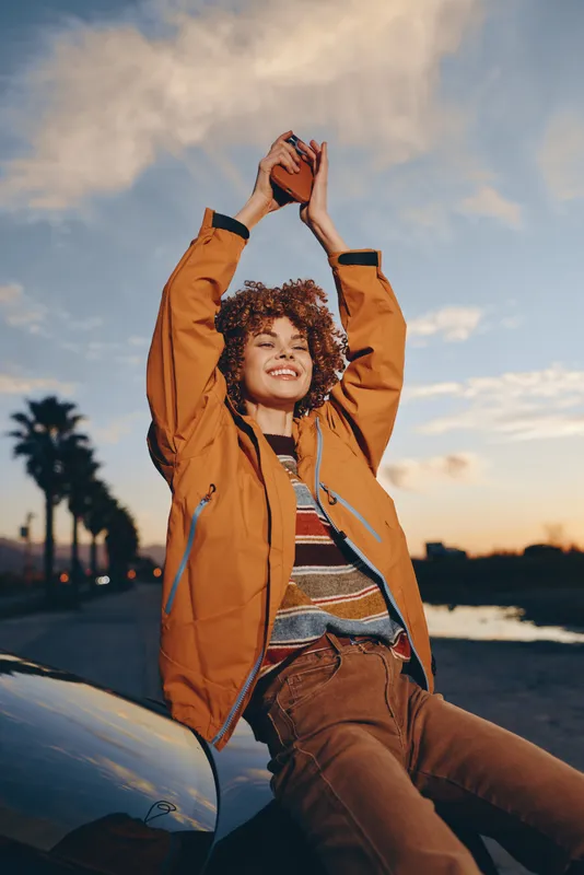 Woman lifestyle car sunset beach jacket smiling outdoors vacation palm setting, young woman sits on car at sunset, tossing hat with joy, warm sky and ocean background, travel mood today
