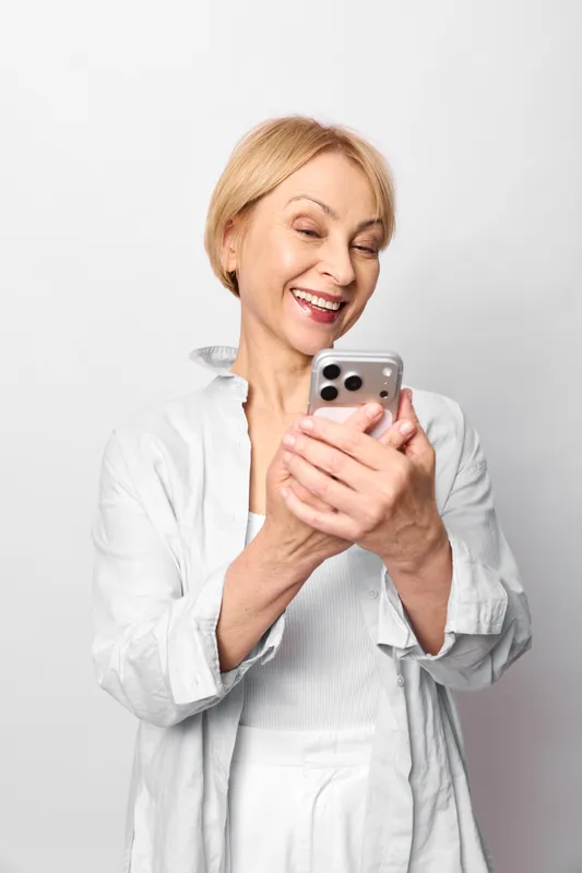Senior woman in medical coat smiles holding smartphone for telemedicine communication using technology to connect with patients calm confident mood in bright clean studio