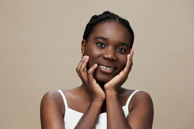 Young Black woman expressing happiness with a natural smile, wearing a minimalist white top, on a soft neutral background, perfect for portraits and lifestyle themes