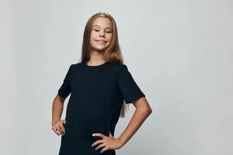 Girl with long hair and black shirt standing confident with hands on hips against plain background. Portrait of happy child with smile and casual style for lifestyle or fashion.