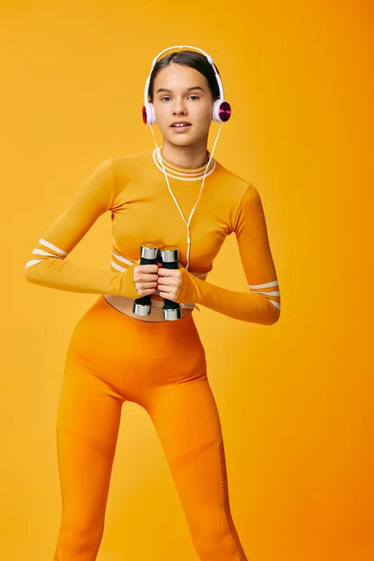 Female fitness enthusiast in bright orange athletic wear holds dumbbells, wearing pink headphones against a vivid orange studio backdrop, conveying energy, focus, and workout momentum.