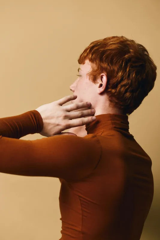 Young man with red hair and fair skin posing in brown turtleneck sweater, side profile with hand near neck against beige background. Fashion, portrait, model and casual style.