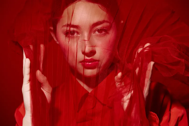 Fashion model behind sheer red fabric holding the material close to her face with intense gaze and minimal makeup. Creative beauty shoot with dramatic red lighting.