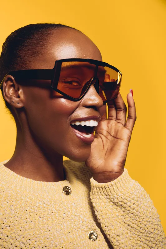 Happy African woman with dark skin wearing oversized sunglasses and a knitted sweater smiles joyfully against yellow background. Concept for fashion, style, and positive emotions.