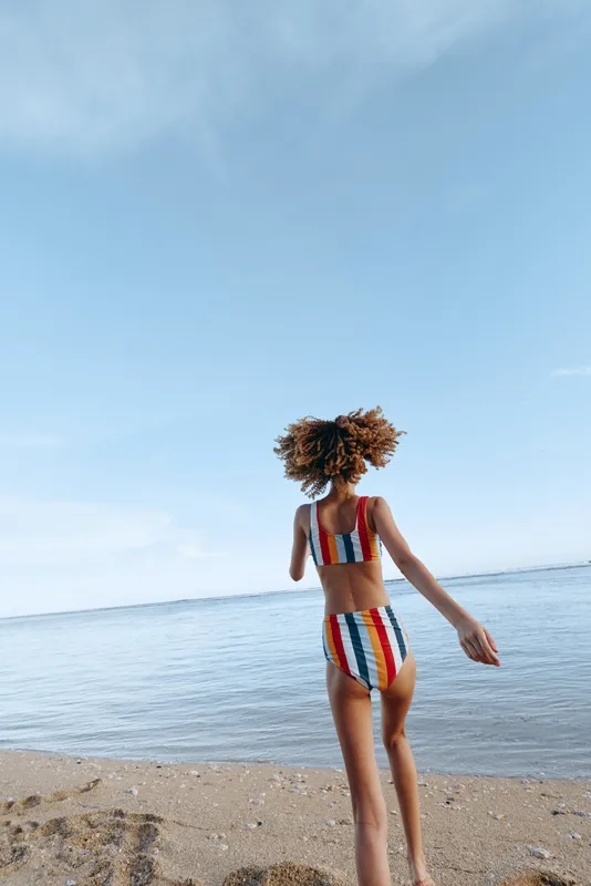Beach ocean swimsuit woman summer striped standing barefoot outdoors sunlight, a young woman in colorful striped bikini walks on sandy shore near calm sea, natural relaxed vacation mood