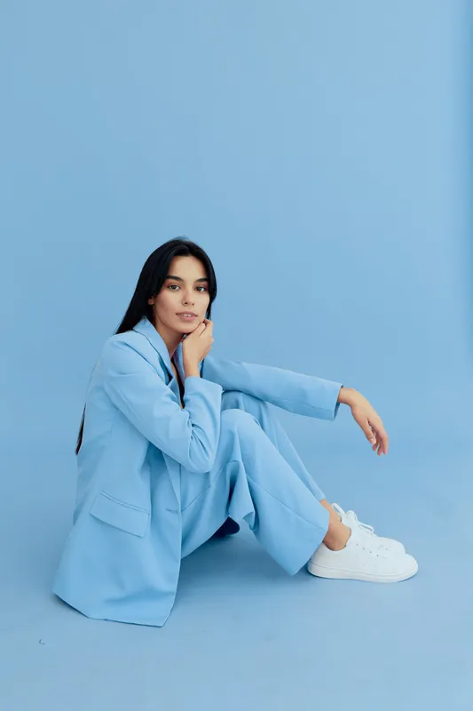 Young woman sitting on floor in pastel blue suit and white sneakers, posing confidently against seamless light blue background for fashion or lifestyle concept.