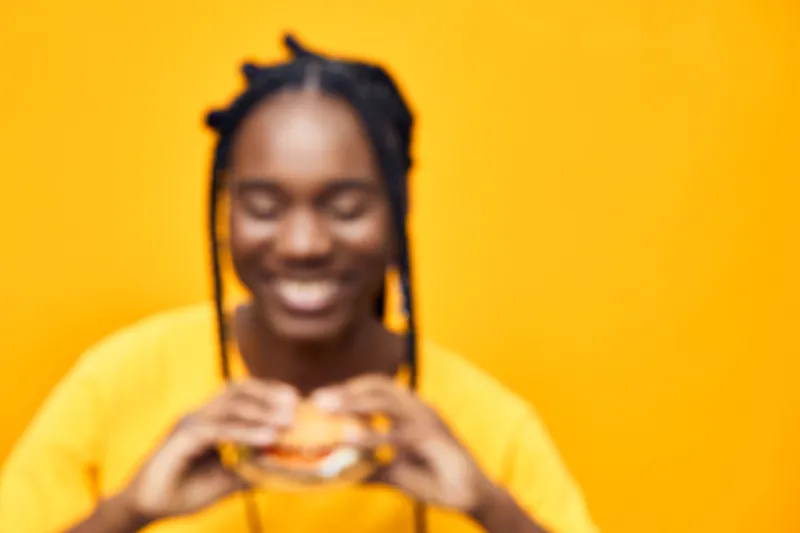 Happy young Black woman enjoying a delicious burger in front of a vibrant yellow background, emphasizing healthy eating and joyful moments