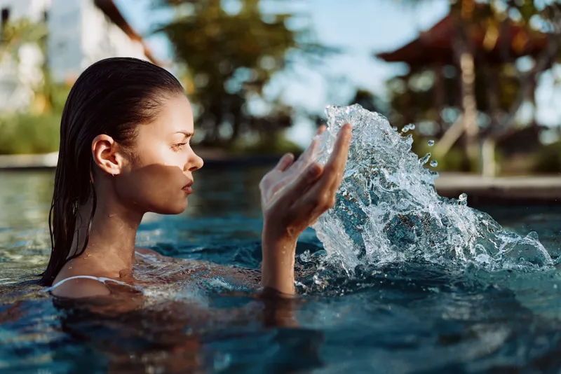 A woman in a swimming pool splashes water with her hands, enjoying a sunny day outdoors. The background features blurred greenery and a warm atmosphere. Concept for lifestyle and relaxation.