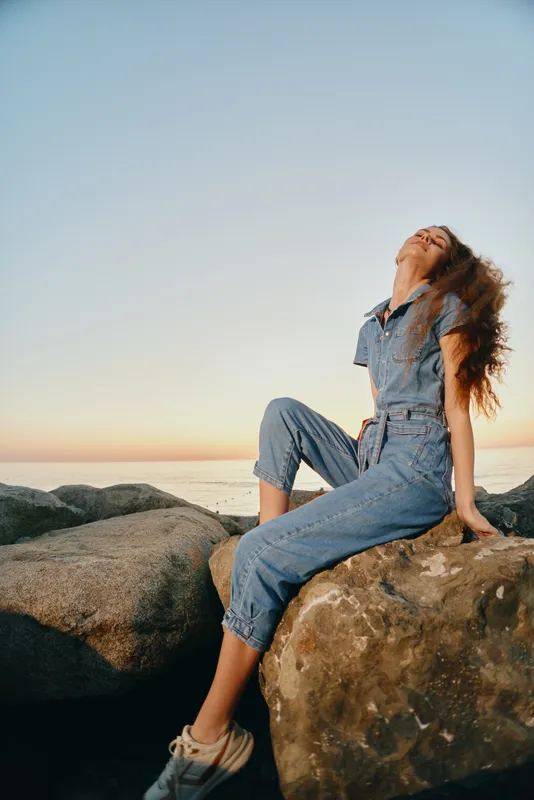 Lifestyle woman in modern boho-western denim street style relaxing on rocks at sunset, film color tones creating warm, natural atmosphere for casual summer mood