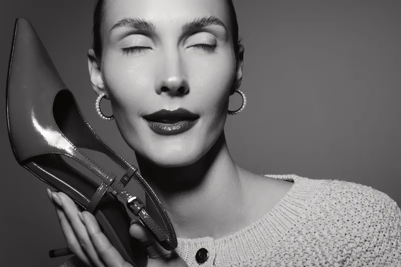 Closeup of white woman with pale skin holding shiny high heel shoe near face, eyes closed, wearing hoop earrings and knitted sweater. Concept of fashion, beauty, and elegance.