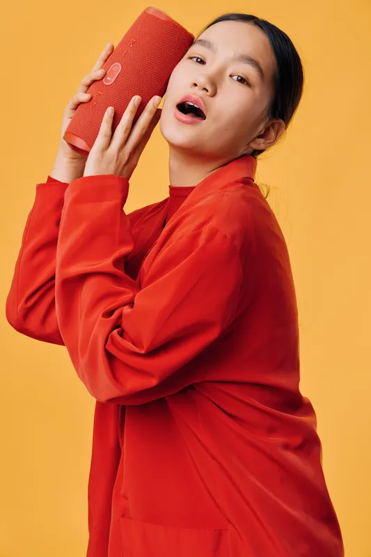 Young woman in red jacket holding portable speaker near ear with open mouth, enjoying music and sound with expressive face on bright yellow background in studio environment.