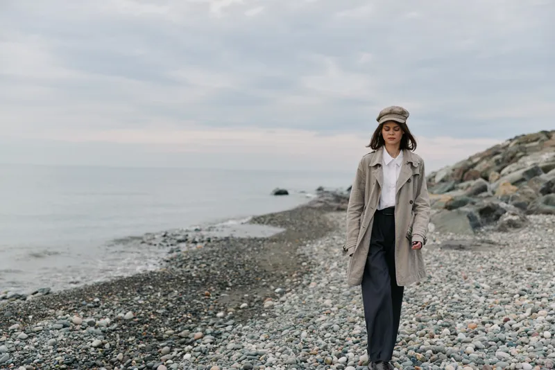 A woman wearing a beige coat and hat walks alone on a rocky beach under a cloudy sky. The calm sea and distant horizon create a peaceful, contemplative mood.