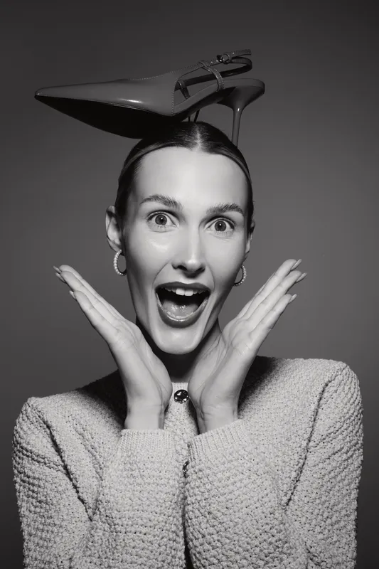 White woman with fair skin wearing high heel shoe on head shows surprised expression with open mouth and hands near face on grey background.