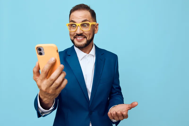 A professional man in a tailored blue suit takes a confident selfie against a solid blue backdrop, expressing optimism, enthusiasm, and approachability with a friendly smile.