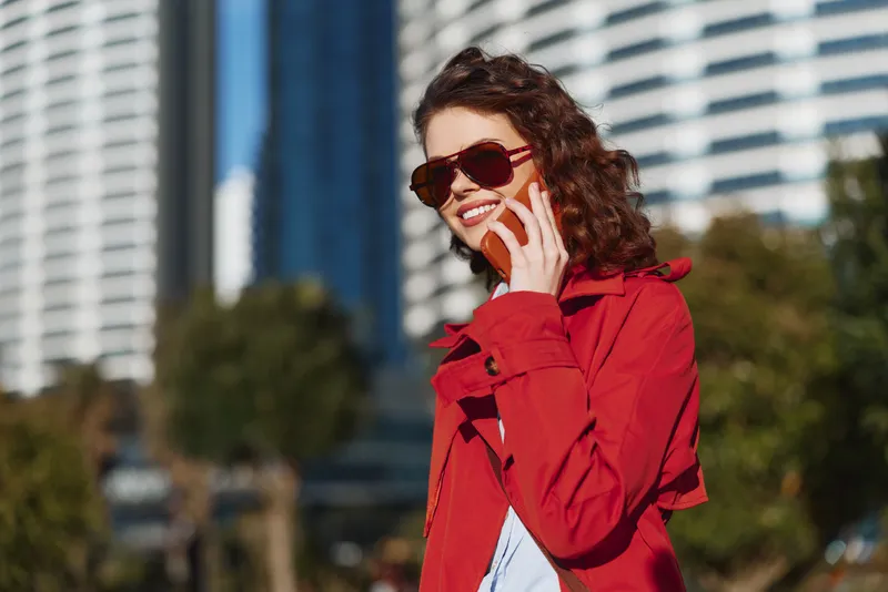 Woman wearing a red coat talks on a smartphone outdoors in an urban setting, smiling behind sunglasses, with modern high rises and blue sky in the background.