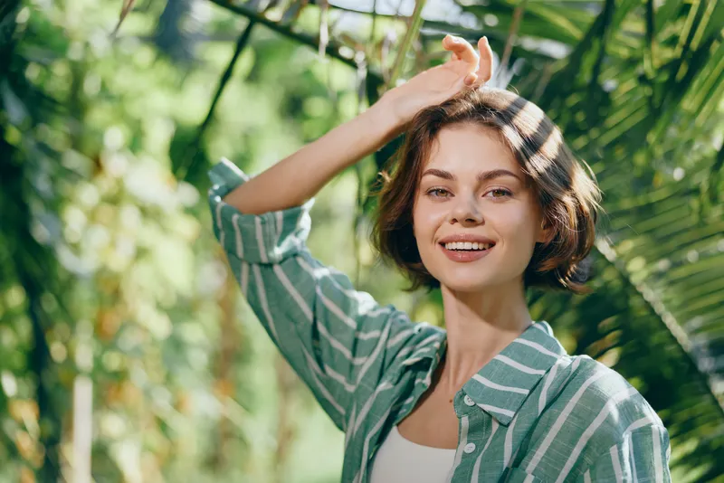 A cheerful woman stands outdoors among lush greenery, sunlight on her face, casual striped shirt, relaxed pose, bright smile, natural portrait conveying joy and confidence in nature.