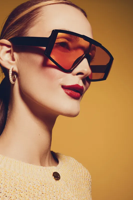 Stylish woman with oversized orange sunglasses and red lipstick poses against yellow background. Fashion portrait with modern accessories and confident expression.