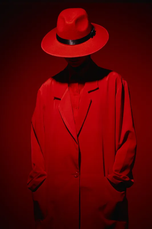 Model in vibrant red hat and matching coat poses under dramatic lighting. Fashion style combined with creative, bold fashion sense in a moody beauty shoot concept.