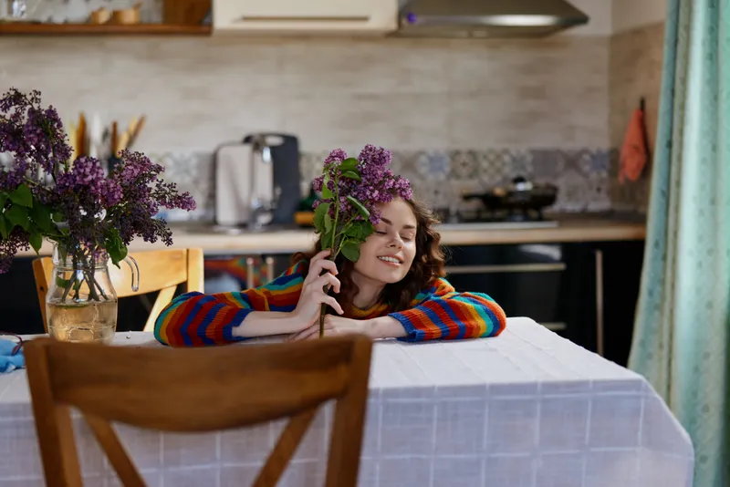 Young woman with curly hair sits at a cozy home table, smiling while holding a lilac bouquet, warm kitchen in the background, soft natural light.