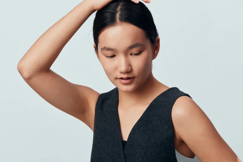 Young Asian woman with black hair wearing sleeveless dark top posing with raised arm and looking down against light gray background in studio for beauty and fashion concept.