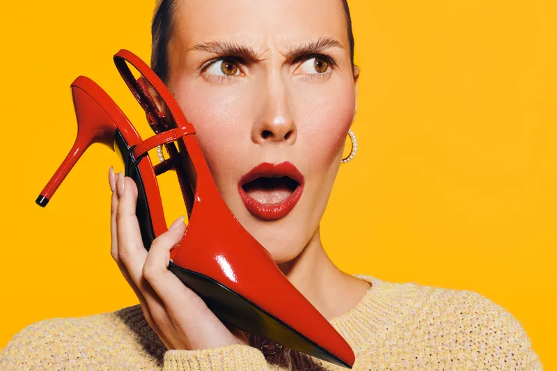 Surprised woman with red lipstick holds red high heel shoe near face on yellow background. Concept of fashion, style, and unexpected emotions in beauty and accessories.
