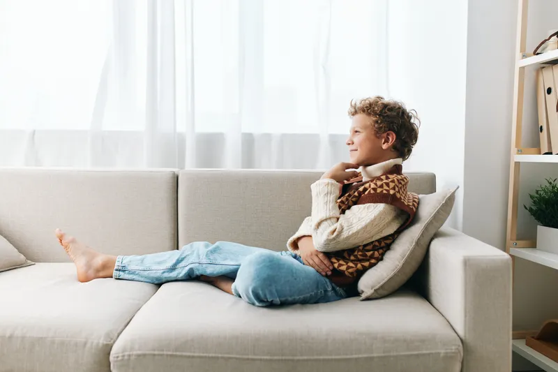 Young boy relaxing on a light sofa, wearing a cozy sweater and scarf, gazing thoughtfully with legs stretched, bathed in soft natural light, a serene domestic moment.