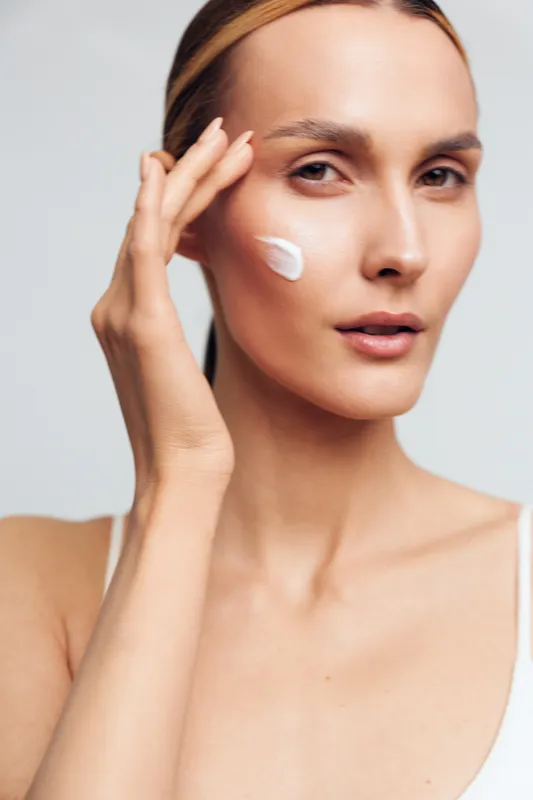 Young woman applying white cream on face, skincare routine, clear skin, natural beauty, close-up portrait, soft lighting, smooth texture, neutral background, healthy lifestyle
