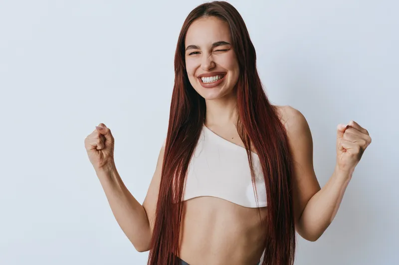 Woman with long hair and fit body celebrating success with clenched fists and smiling face. Female, happy and confident in casual top on plain background for positivity.